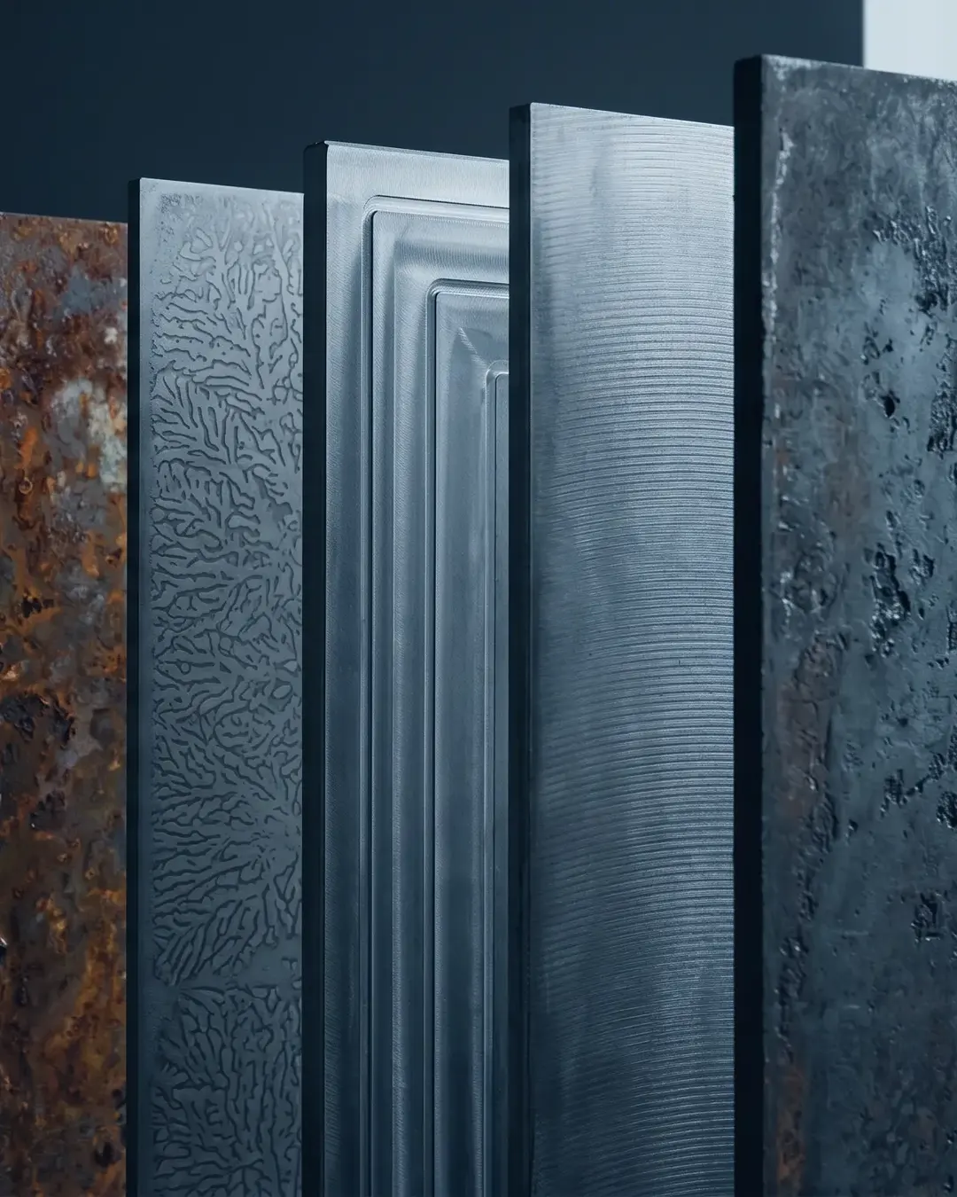 Upright metal slabs side by side in cold light, each a different surface from corroded to etched, machined, brushed and cast, evoking the six service pillars