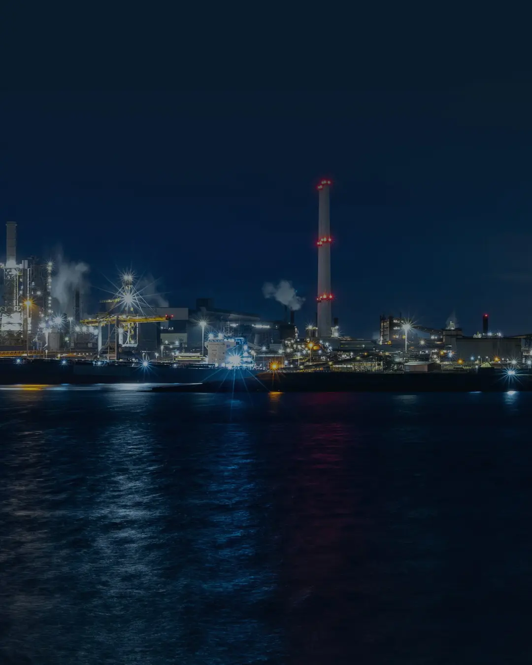 The waterfront of a process plant at night, lit columns and a flare stack reflected in dark water