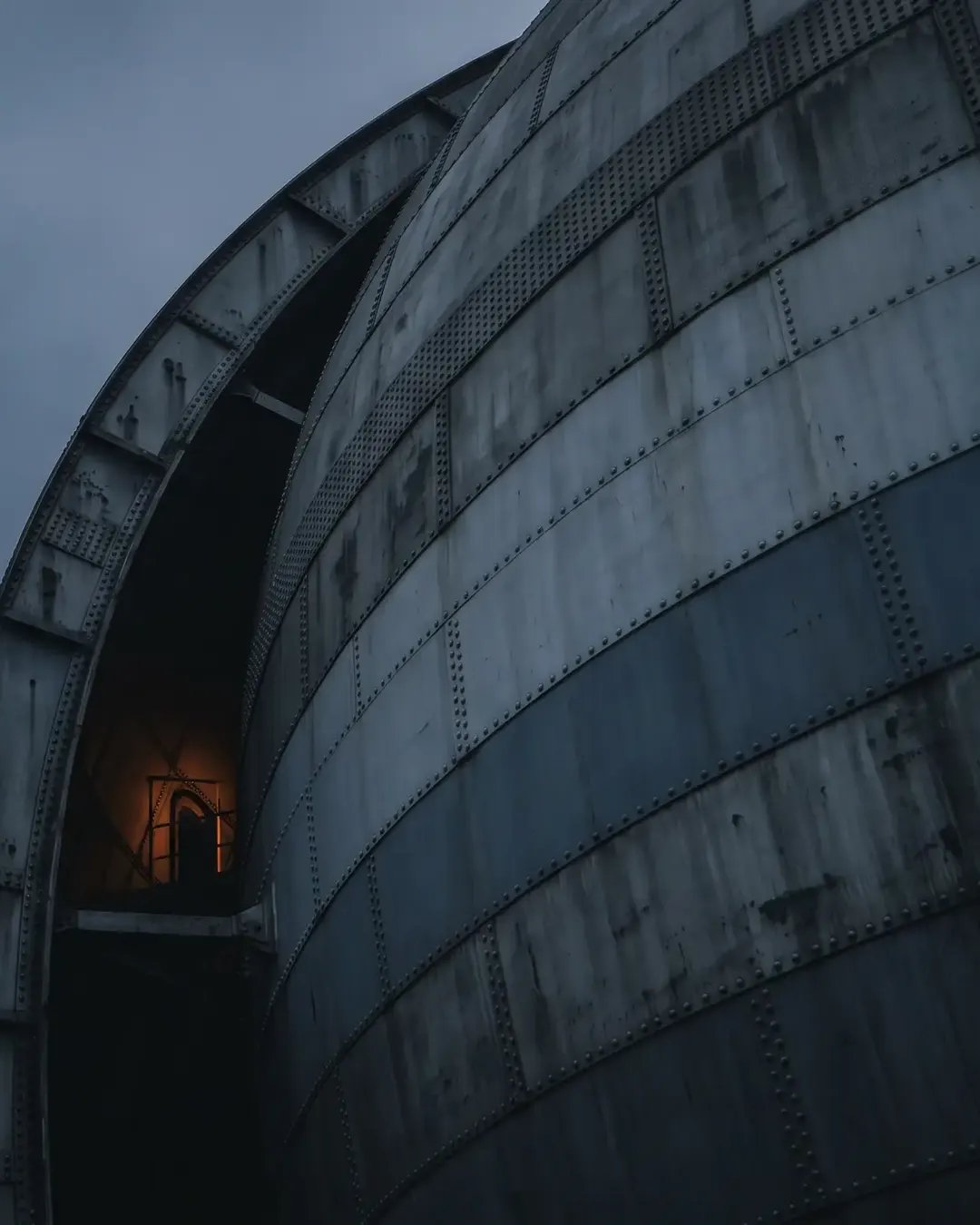 The curved shell of an enormous pressure vessel seen from below at an oblique angle, cold dusk sky to the left
