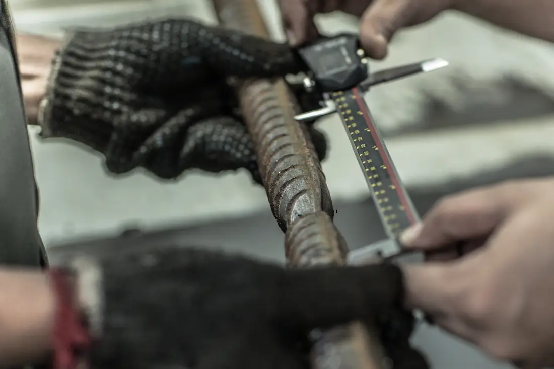 A digital vernier caliper measuring across the ribs of a deformed reinforcement bar, the bar steadied in a gloved hand