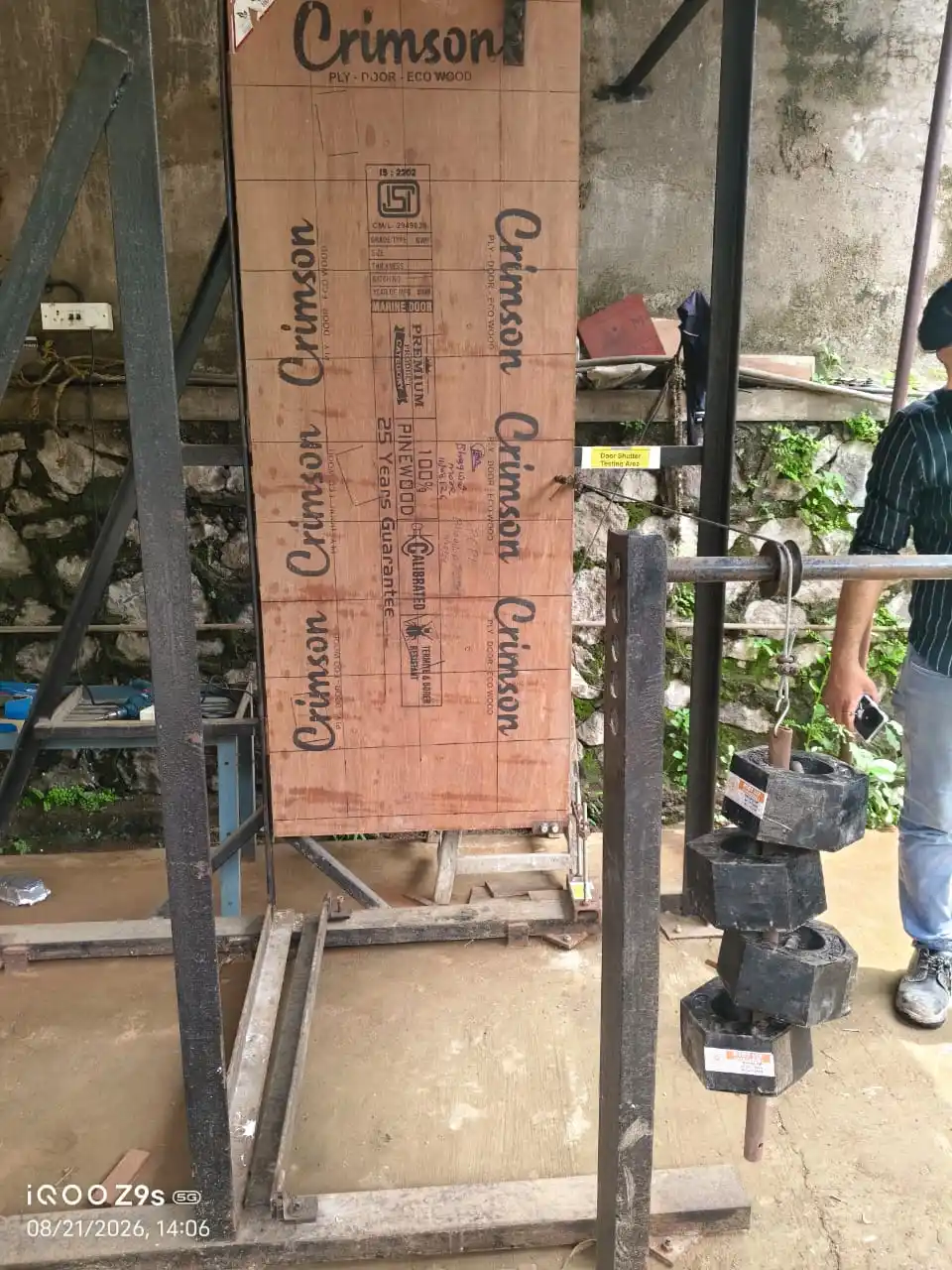 A flush door shutter clamped in a test frame with a stack of calibrated weights hanging from a bar at its edge