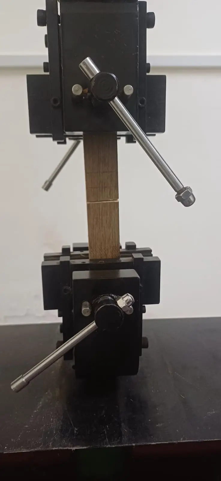 A short square plywood specimen held between two grips, the veneer laminations visible along its side