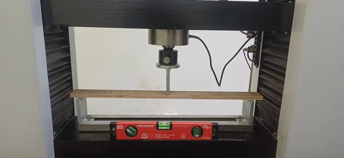 A plywood strip on two supports in a testing machine, a loading nose bearing on its centre, a spirit level set on the frame beneath