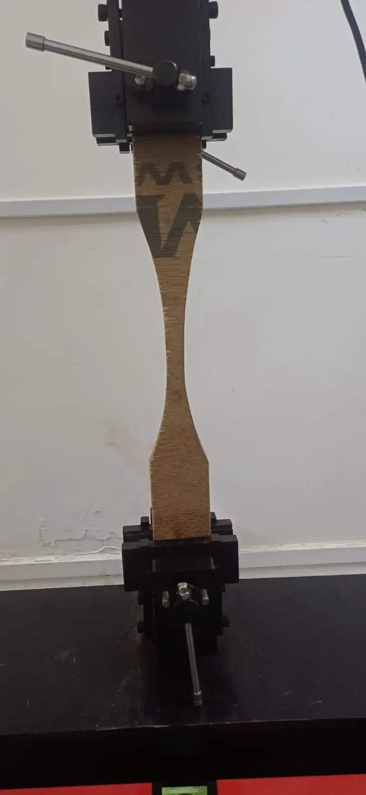 A waisted plywood tensile specimen clamped in the upper and lower grips of a testing machine