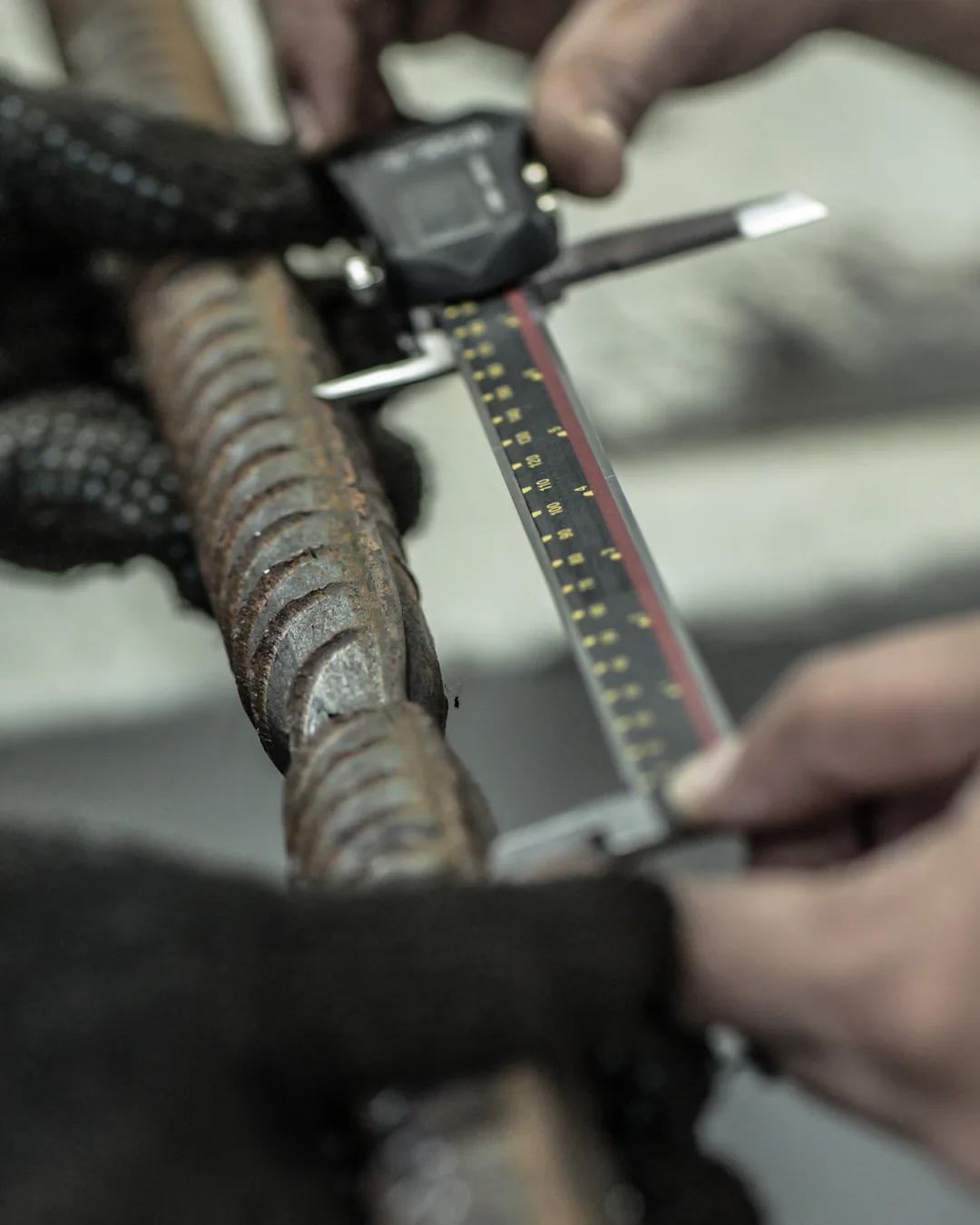 A digital vernier caliper measuring across the ribs of a deformed reinforcement bar, the bar steadied in a gloved hand