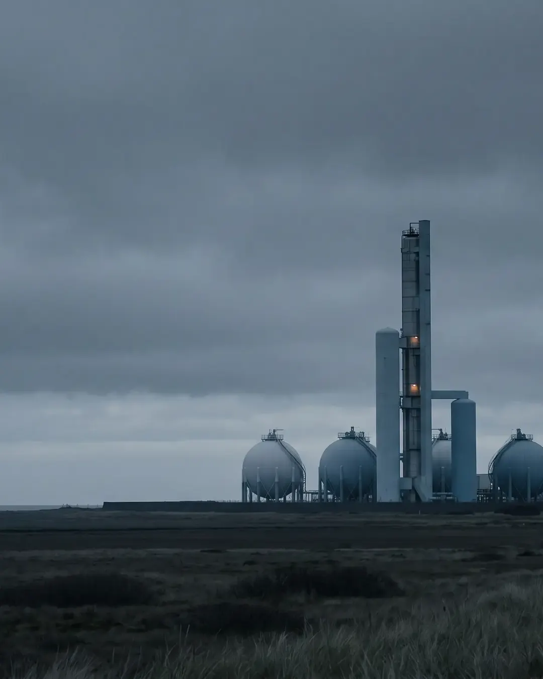 Ammonia storage spheres and a prilling tower on a flat coastal plain under a heavy grey sky