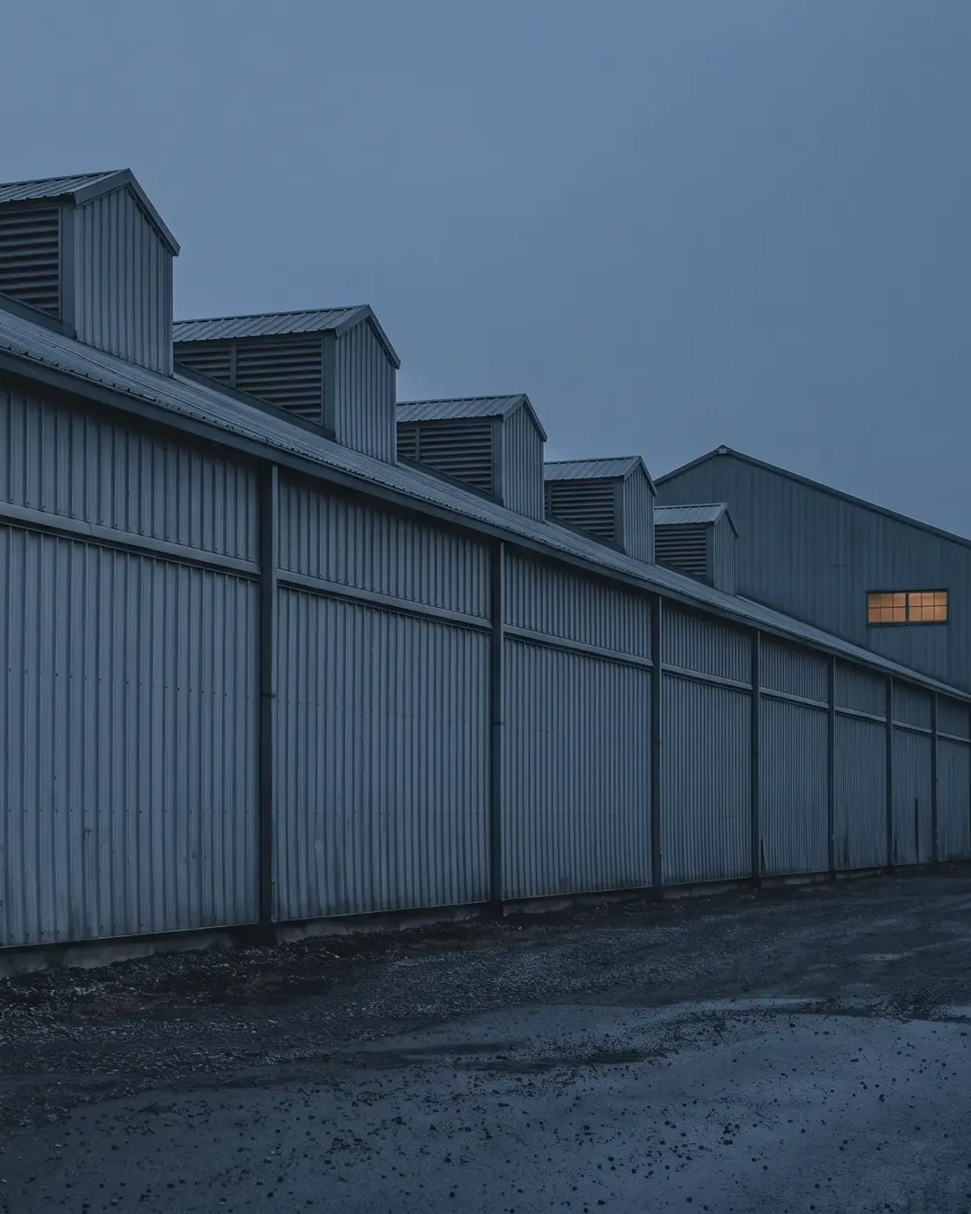 The exterior of a long rolling-mill shed at dusk, ribbed steel cladding receding to a vanishing point