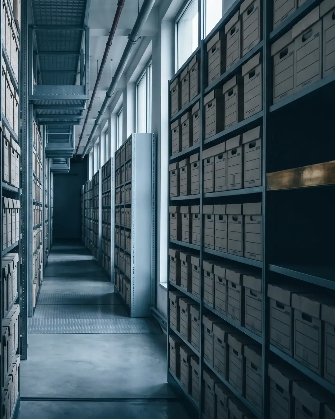 A long, dim archive corridor of grey steel shelving holding uniform document boxes