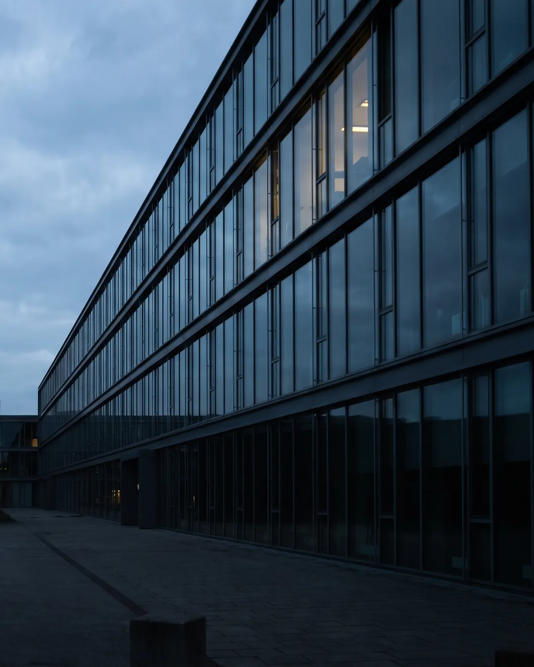 The glazed facade of an anonymous industrial laboratory building at blue hour, a few upper windows lit, empty forecourt in the foreground