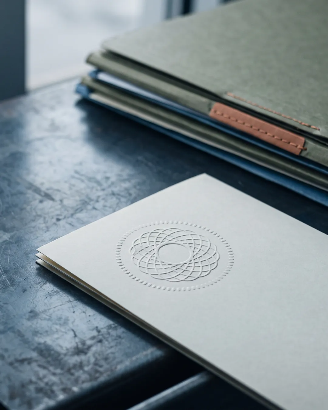 A heavy embossed seal impression in thick cream paper on a dark steel surface, with closed document folders falling out of focus behind it