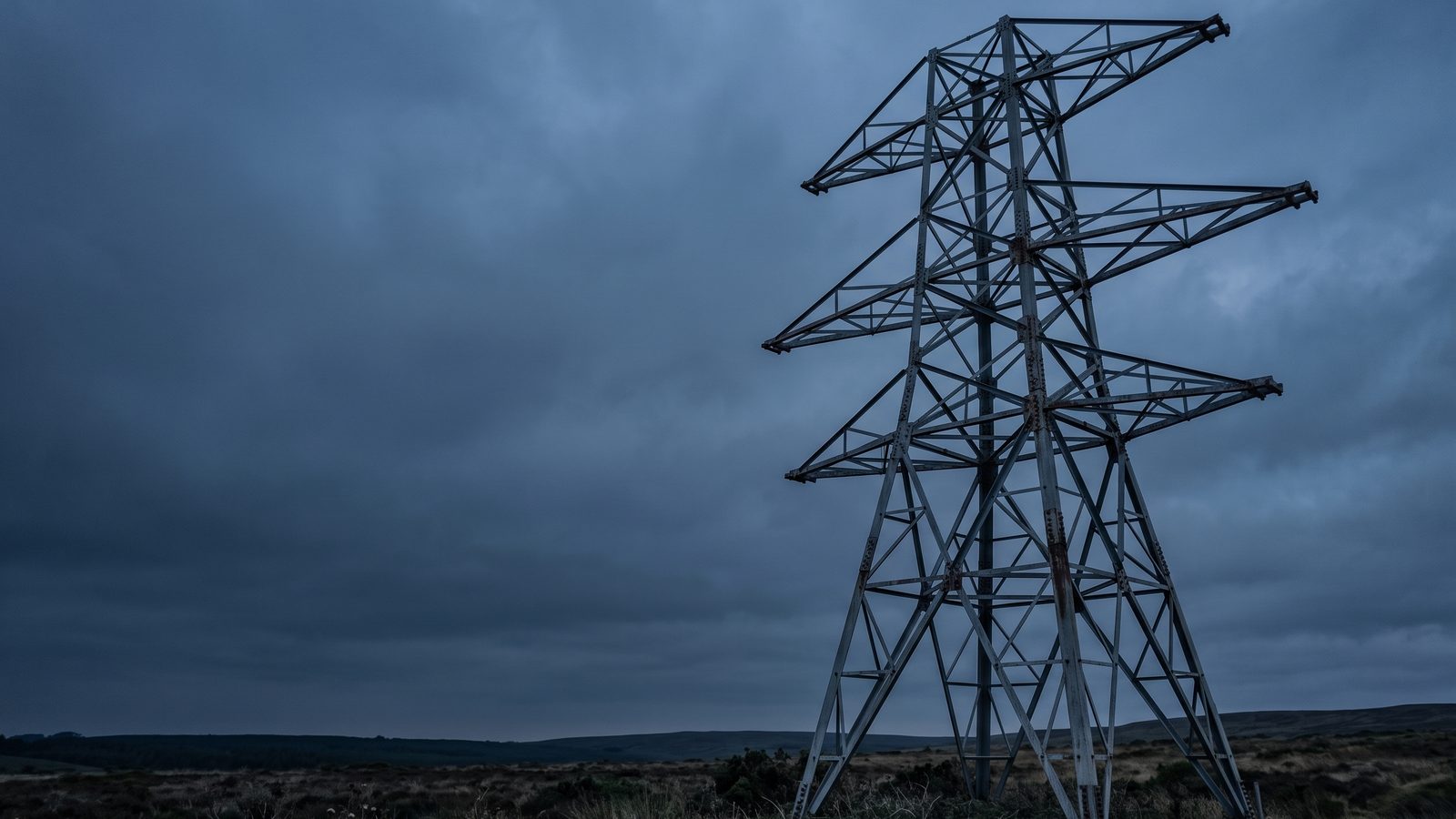 A weathered galvanised steel lattice transmission tower with three crossarms standing in open scrubland under an overcast evening sky