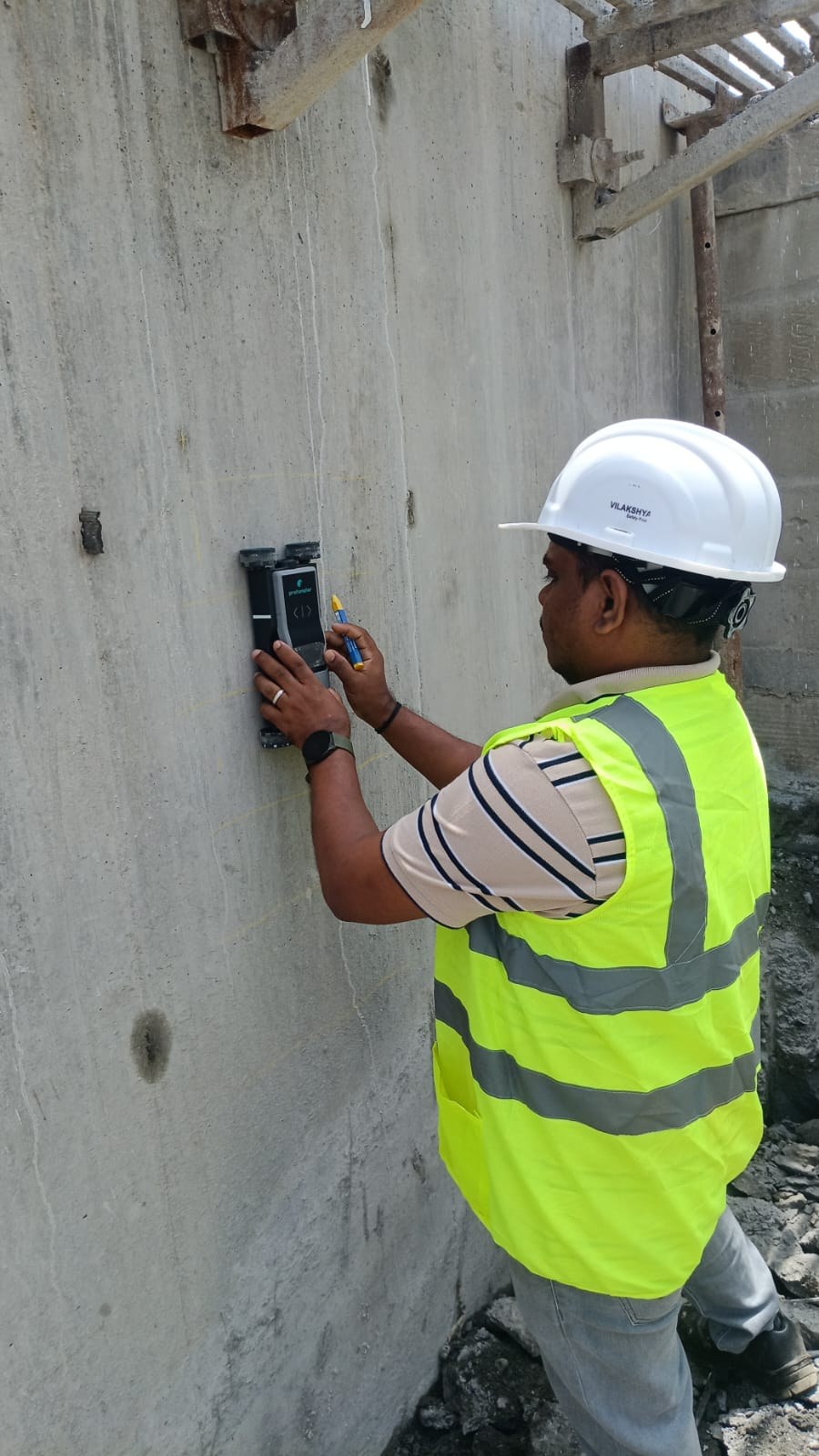 A TCR engineer in a hard hat and high-visibility vest holding a cover meter against a concrete wall marked out with a chalk grid