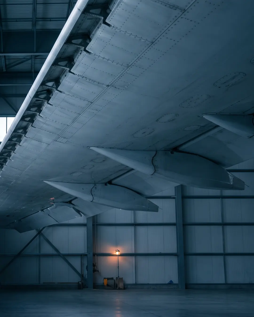 The underside of a large aircraft wing receding through a dark assembly hall, its skin panels and structural ribs catching cold diffuse light